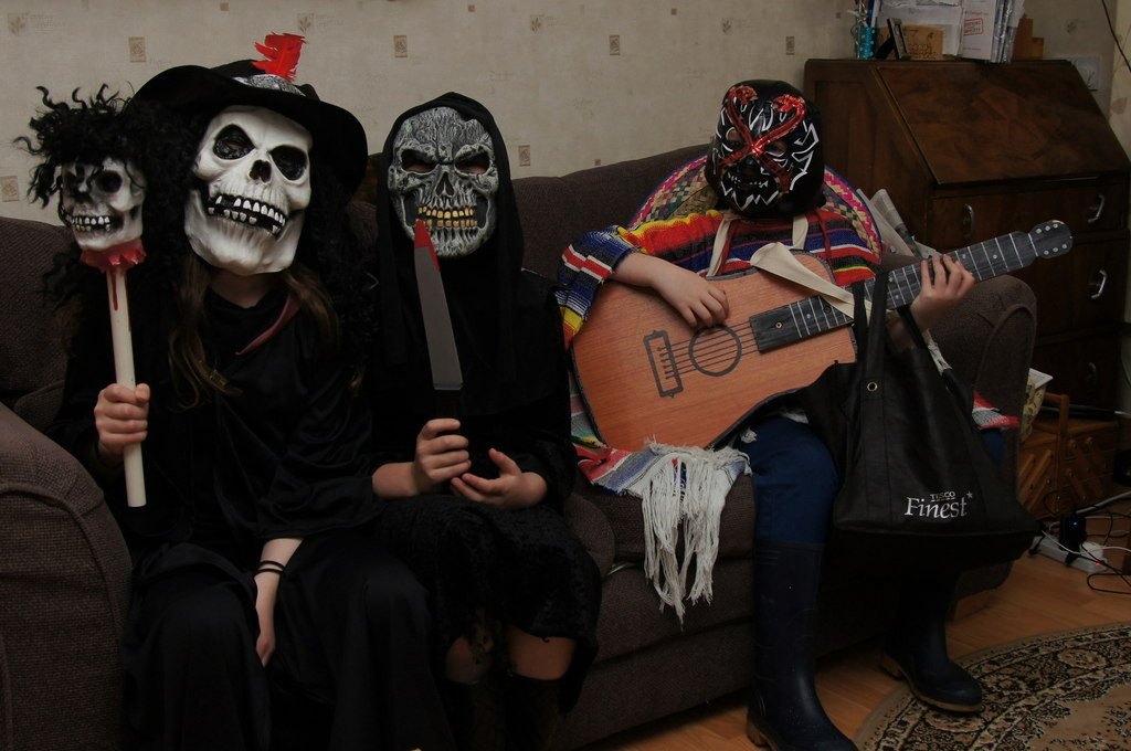 Four Halloween guisers in skull and wrestling masks, one holding a toy guitar, sitting on a sofa in Baltasound, Shetland