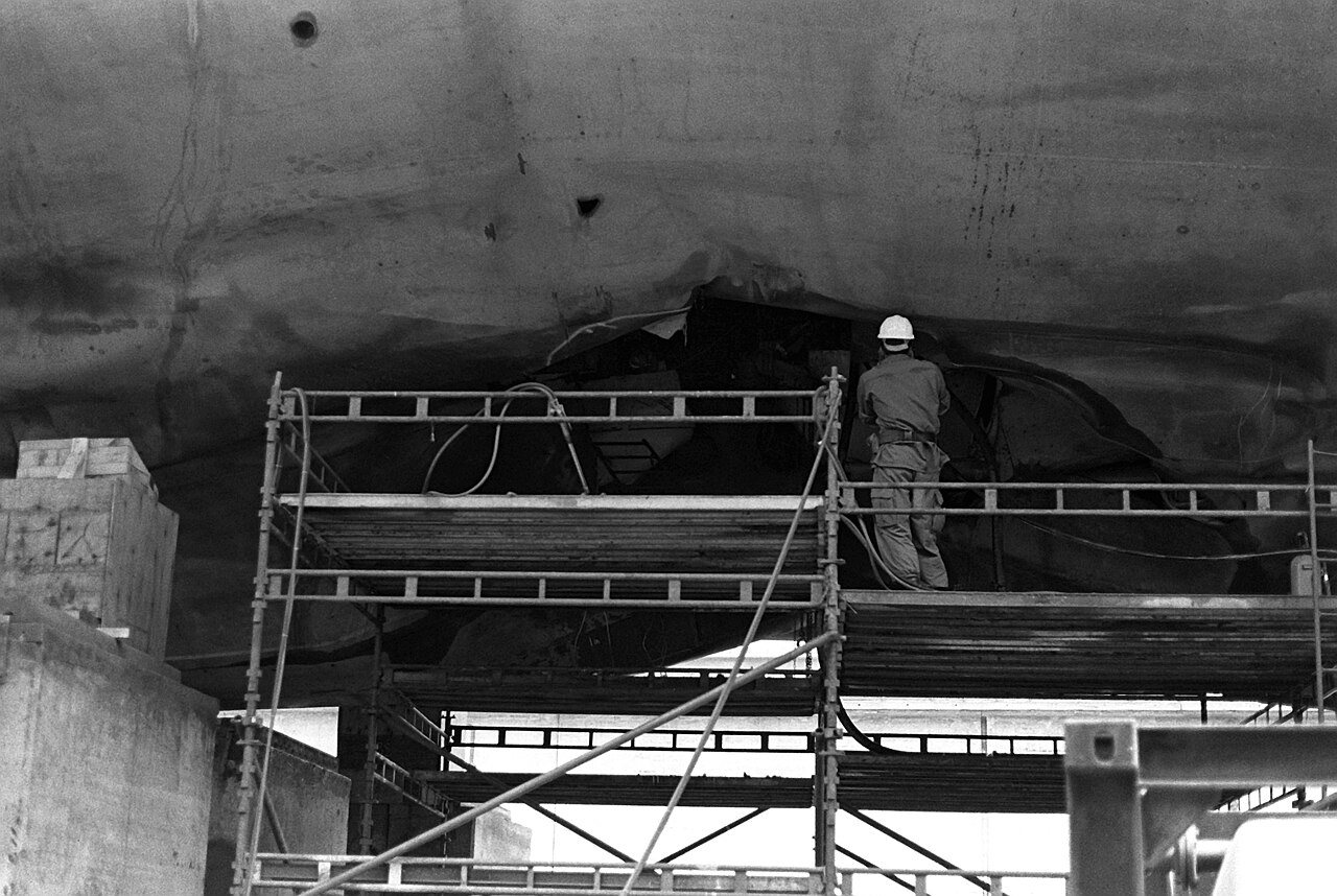 Black-and-white photograph of a worker in a hard hat on scaffolding in dry dock, inspecting the buckled hull of the frigate USS Samuel B. Roberts after it struck an Iranian mine in April 1988